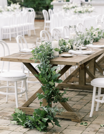 Outdoor wooden table set for an event, adorned with greenery and white place settings. White chairs surround the table on a stone-paved area.